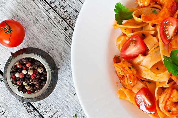 A dish of pasta on a table accompanied by a bowl of pepper corns and a whole tomatoe