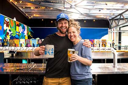 Smiling couple holding up glasses of beer behind the bar in a brewery Mountain Culture Beer Co bar