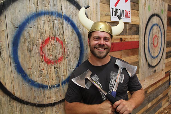 Man holding 2 axes standing in front of wooden targets