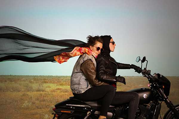 Two women on black motorbike with large black scarf blowing in wind behind them