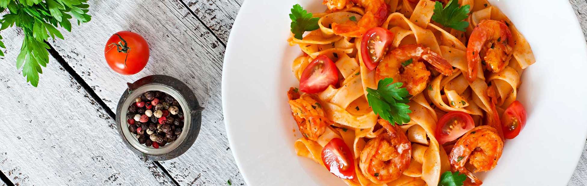 A dish of pasta on a table accompanied by a bowl of pepper corns and a whole tomatoe
