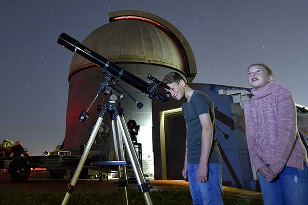 Boy looking through telescope, girl looking at sky with observatory behind them