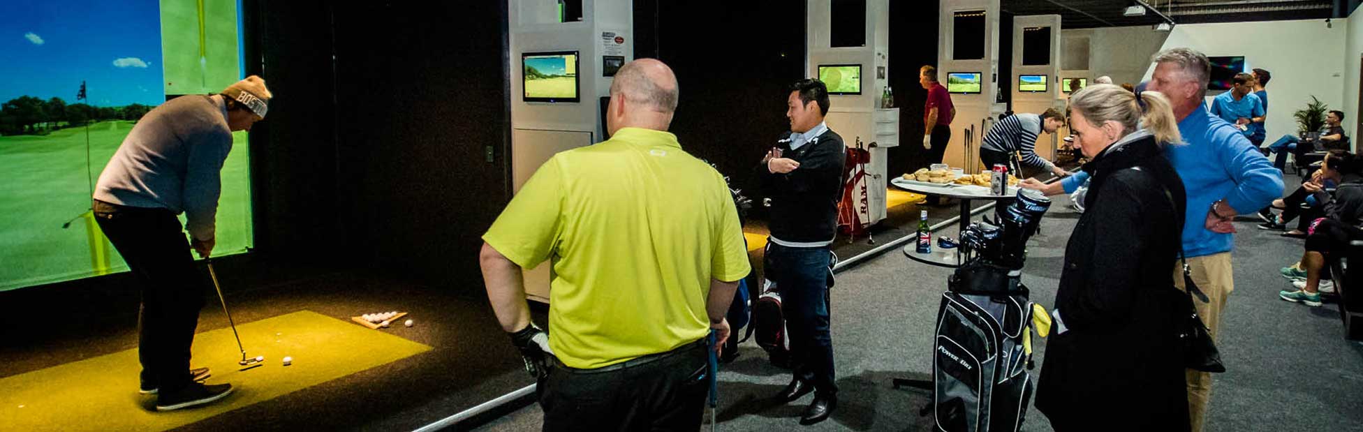 A group of people standing around watching a man prepare to swing at a golf ball indoors in front of a screen