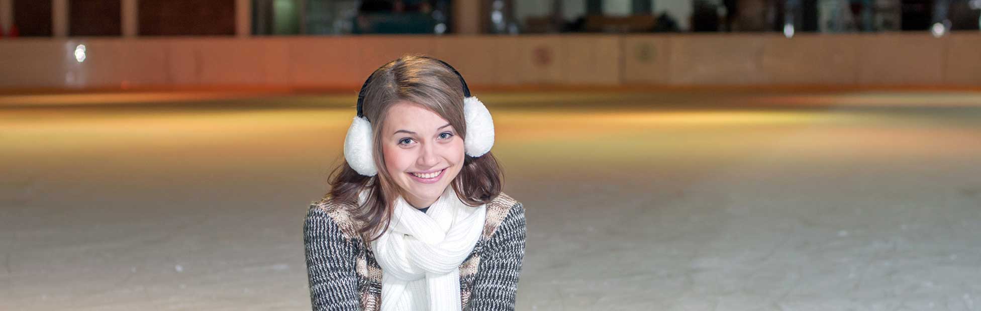 Young girl sitting on an ice rink wearing ear muffs, a scarf to stay warm